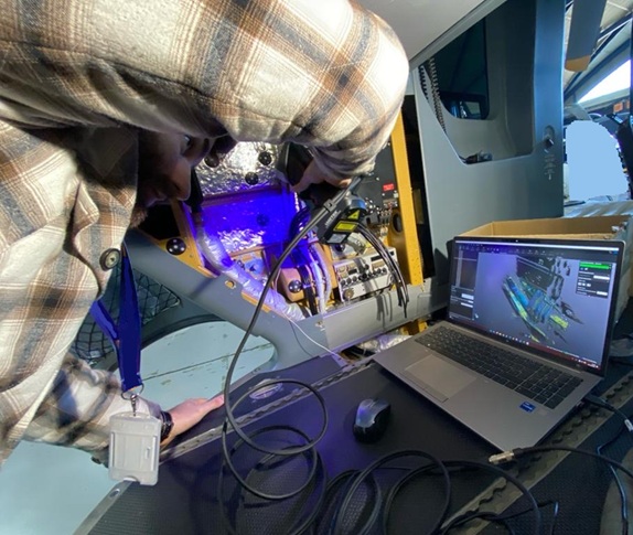 Engineer using a handheld 3D scanner to capture the interior structure of an aircraft cockpit, while real-time scan data appears on a laptop screen running 3D metrology software. 